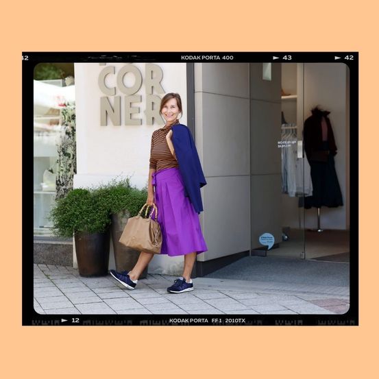 Eine Frau in einem lila Rock und einer blauen Jacke geht vor einem Geschäft. Ein Schild sagt 'COR NER'. Töpfe mit Pflanzen und Taschen sind in der Nähe.