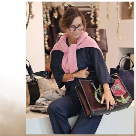 Eine Frau mit Brille und einem rosa Schal sitzt auf einer Bank in einem Bekleidungsgeschäft und hält eine braune Tasche mit Blumendesign.