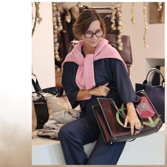 Eine Frau mit Brille und einem rosa Schal sitzt auf einer Bank in einem Bekleidungsgeschäft und hält eine braune Tasche mit Blumendesign.