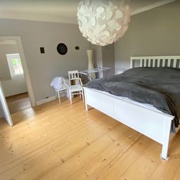 Ein Schlafzimmer mit Holzboden, einem weißen Bett und einer weißen Laterne. Es gibt zwei weiße Stühle und einen Tisch mit einem Metallständer. Der Raum hat eine graue Wand und ein Fenster.