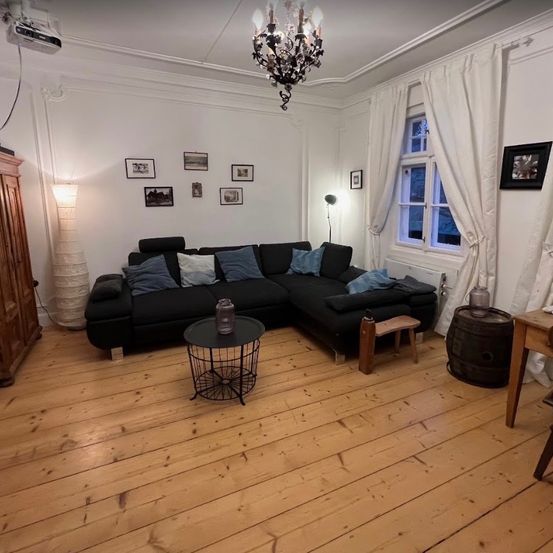 Ein Wohnzimmer mit einem schwarzen Ecksofa, Holzboden und weißen Wänden. Eine Kronleuchter hängt von der Decke, und ein Fenster mit weißen Vorhängen befindet sich rechts.