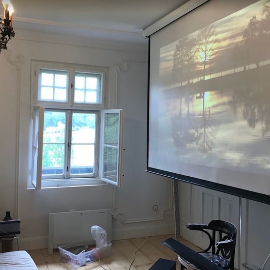 Ein Zimmer mit einer Projektionsleinwand, die einen Sonnenuntergang über Wasser zeigt, einem offenen Fenster und einem Heizkörper darunter.