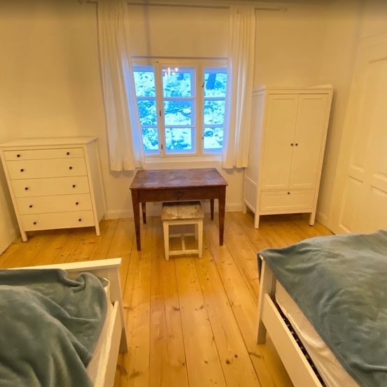 Ein Schlafzimmer mit zwei Betten, einem Holztisch und einem kleinen Hocker vor einem Fenster mit weißen Vorhängen. Es gibt zwei weiße Kommoden auf beiden Seiten des Raumes.