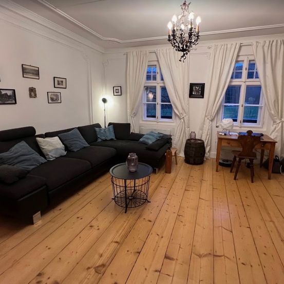 Ein Wohnzimmer mit einem schwarzen Ecksofa, einem Holzboden und weißen Wänden. Es gibt einen Kronleuchter, einen Schreibtisch und einen Fass auf dem Boden.