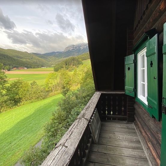 Ein Balkonblick auf ein Holzhaus mit grünen Fensterläden, mit Blick auf ein saftiges grünes Feld und entfernte Berge unter einem bewölkten Himmel.