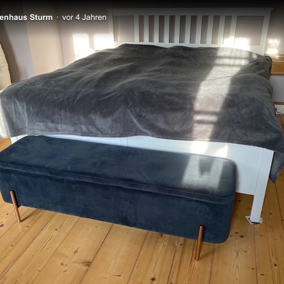 Ein blauer Samtbank mit Holzbeinen steht vor einem Bett mit einer grauen Decke. Das Bett steht an einer Wand mit einem Holzregal darüber.