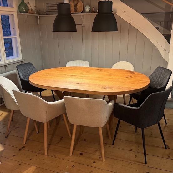 Ein Esszimmer mit einem runden Holztisch, umgeben von sechs Stühlen, zwei schwarz und vier beige, vor einer weißen Wand mit Fenster und hängenden Lampen.