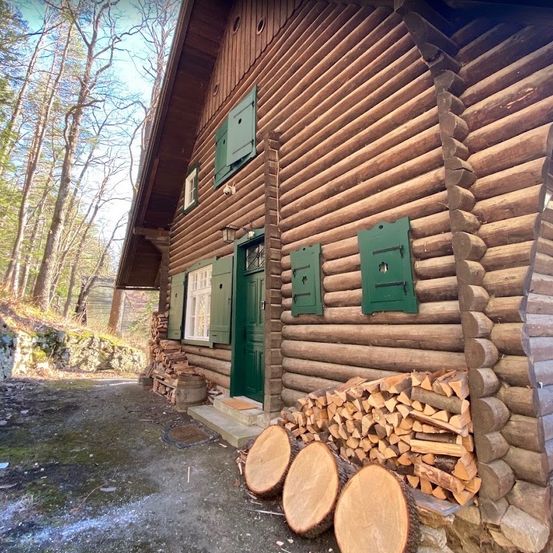 Ein Holzblockhaus mit grünen Fensterläden und einer grünen Tür, umgeben von einem Haufen Feuerholz und Baumstämmen.