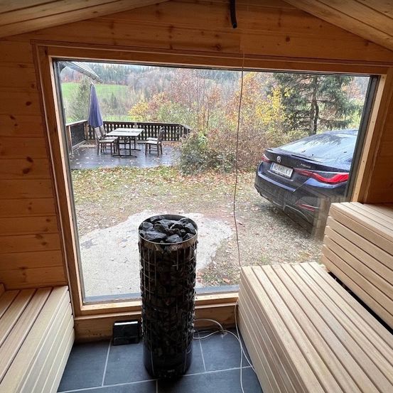 Ein Blick aus einem Holzsauna zeigt eine Terrasse mit Tisch, Stühlen und einem Regenschirm, ein geparktes Auto draußen und einen Holzofen.