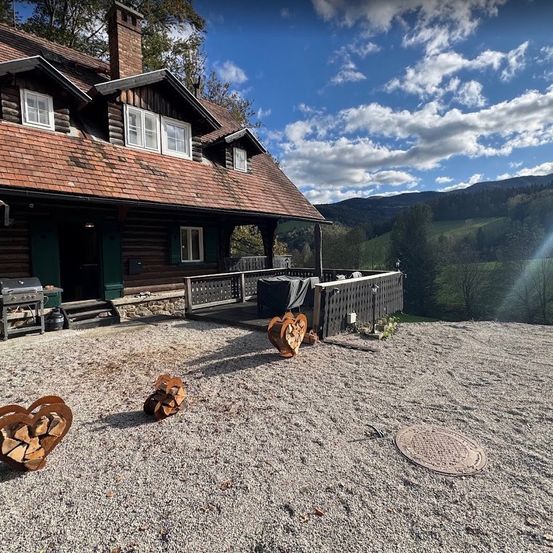 Ein Blockhaus mit braunem Dach und weißen Fenstern steht in einem Kieshof, mit einem Bergrücken im Hintergrund. Herzförmige Holzstapel und ein Grill sind vor dem Haus.