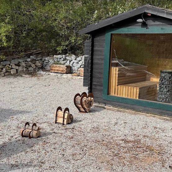 Eine kleine Holzhütte mit grünem Rahmen und Glasfenster, umgeben von einem Kiesbereich mit herzförmigen Holzhaltern und einer Steinmauer im Hintergrund.