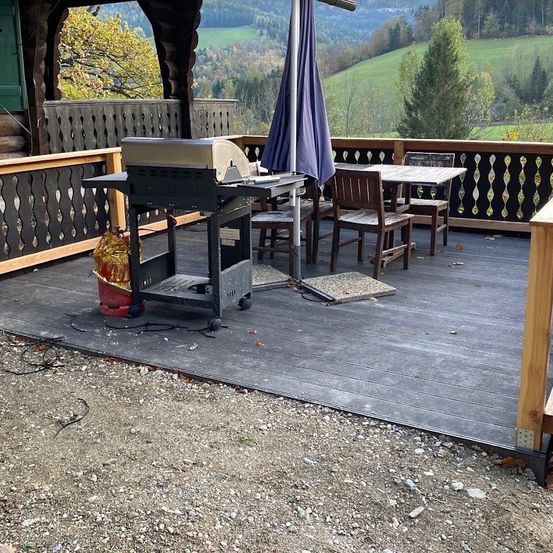 Eine Holzterrasse mit einem Grill, einem Tisch und Stühlen sowie einem geschlossenen Regenschirm. Die Terrasse ist von einem Holzzaun umgeben. Im Hintergrund befindet sich ein grünes Feld mit Bäumen.