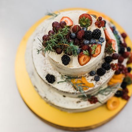 Ein dreistöckiger Kuchen mit weißer Glasur, belegt mit verschiedenen Früchten wie Erdbeeren, Blaubeeren und Brombeeren, verziert mit grünen Blättern auf einem gelben Teller.
