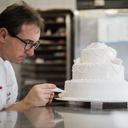 Ein Mann in einer Kochuniform dekoriert eine dreistöckige weiße Hochzeitstorte. Er verwendet eine Spritztüte, um feine Details hinzuzufügen. Der Schauplatz scheint eine Bäckerei zu sein.