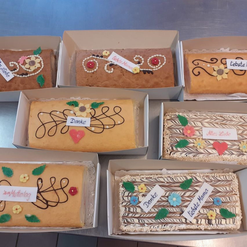 Eine Reihe von sechs rechteckigen Kuchen in Schachteln, jeder mit einzigartigen Dekorationen und Etiketten wie 'Danke', 'Muttertag' und 'Beste Mama'.