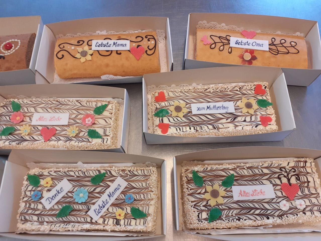 Fünf dekorierte Kuchen in Pappkartons, jeder mit personalisierten Botschaften wie 'Liebste Mama' und 'Alles Liebe'. Sie sind mit Blumen, Herzen und Schokoladendesigns verziert.