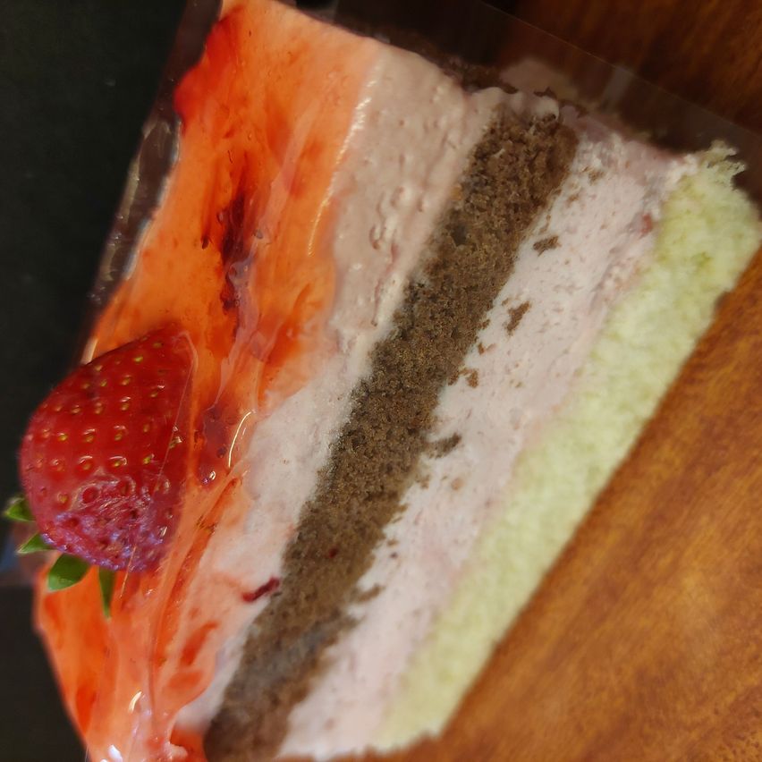 Ein Stück geschichteter Nachtisch mit Erdbeere obenauf, in Plastik verpackt auf einer Holzoberfläche.