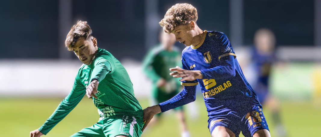 Zwei Fußballspieler, einer in Grün und einer in Blau, befinden sich in einer dynamischen Aktion auf einem Feld. Der Spieler in Grün tritt den Ball, während der Spieler in Blau versucht, ihn zu blockieren. Das Event ist für Samstag, den 14. Februar um 18 Uhr geplant.