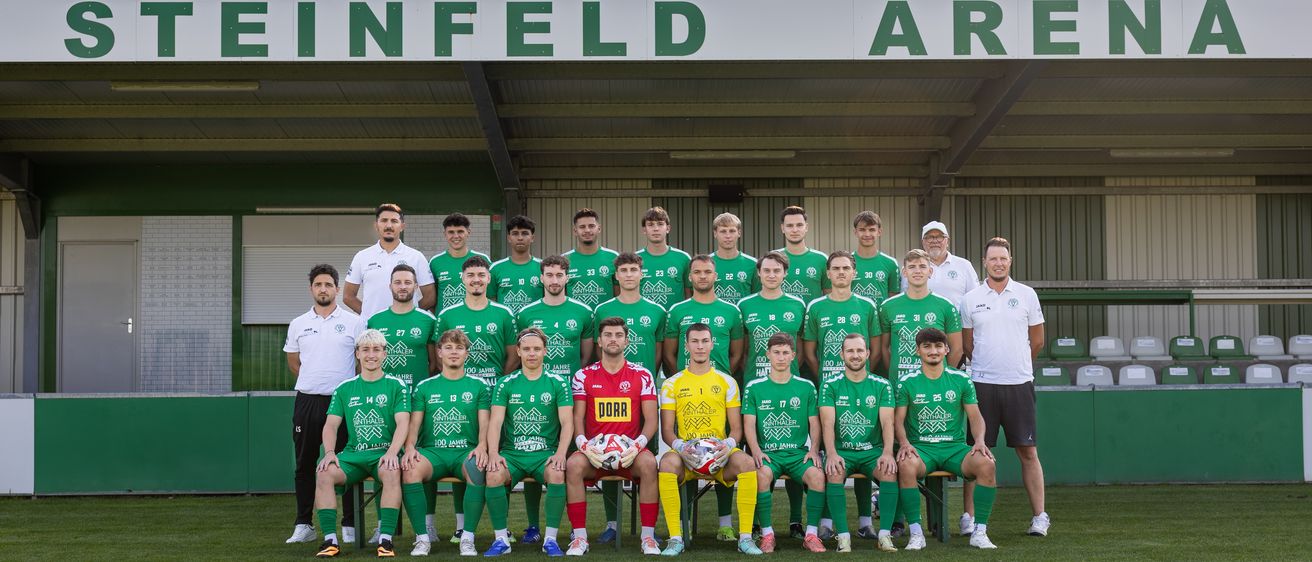 Eine Fußballmannschaft posiert für ein Foto auf einem grünen Rasen mit einem weißen Banner mit der Aufschrift 'EINFELD ARENA' im Hintergrund.