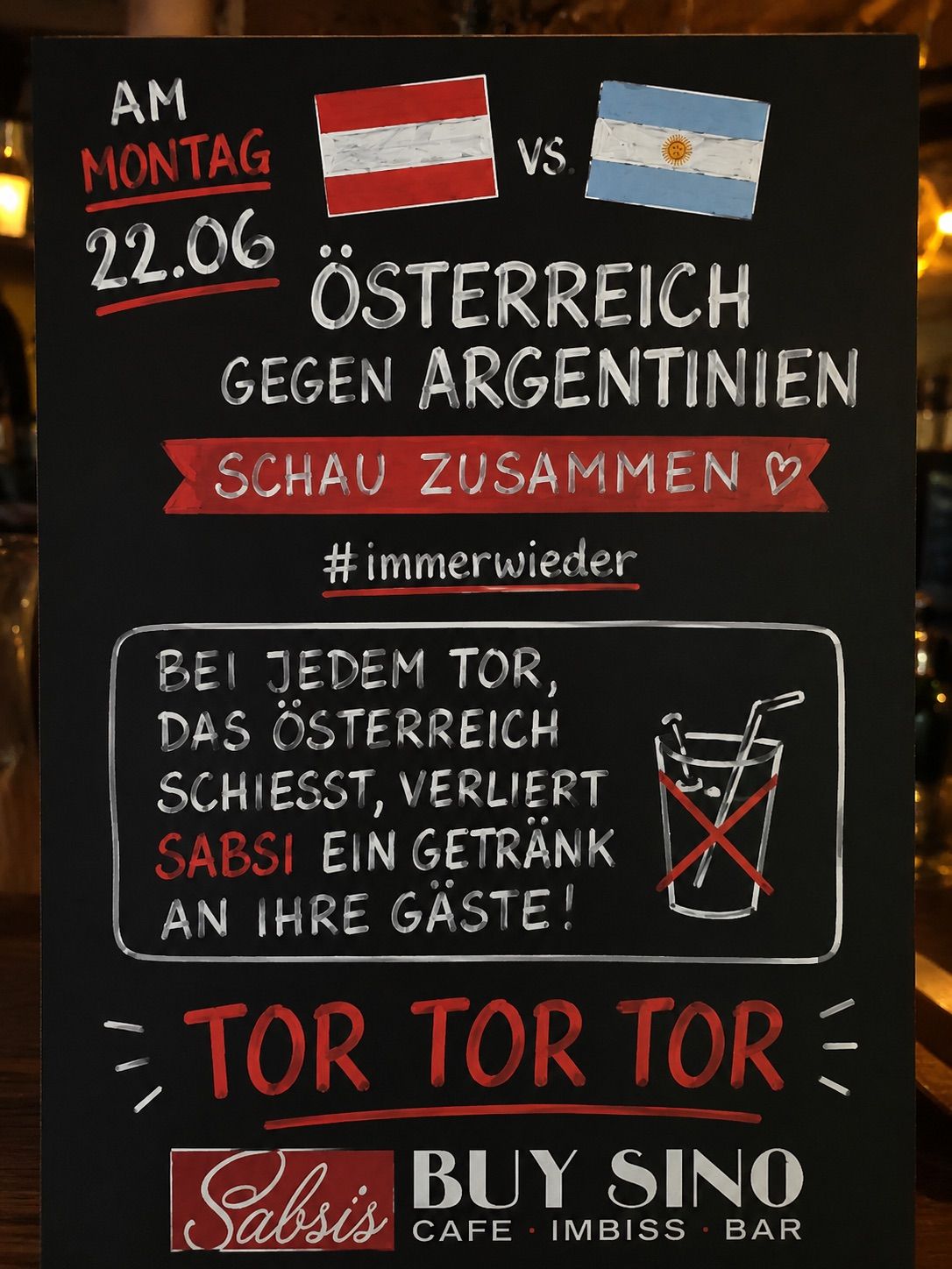 Ein Schwarzes Brett mit einer Fußballspiel-Ankündigung. Das Datum ist 22.06. Österreich spielt gegen Argentinien. Der Slogan ermutigt alle, gemeinsam zuschauen. Es gibt auch eine Nachricht, die besagt, dass Gäste bei jedem Tor Österreichs ein Getränk verlieren. Das Brett endet mit dem Wort 'Tor' dreimal wiederholt.