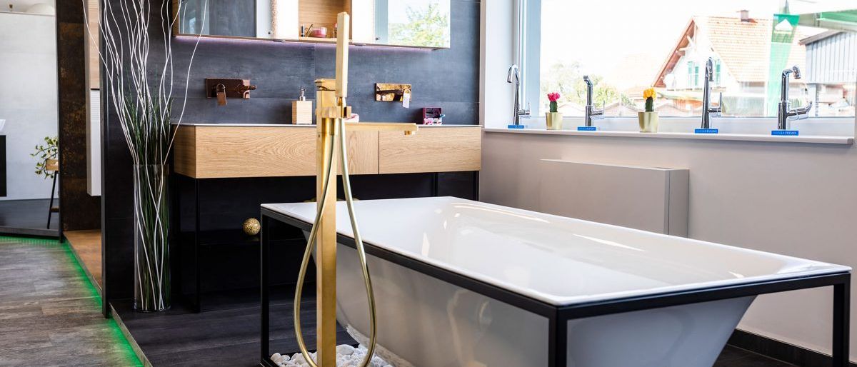 Ein modernes Badezimmer mit einer weißen Badewanne und goldenem Wasserhahn, Holzschränken und einem großen Spiegel, der das Äußere mit Pflanzen reflektiert.