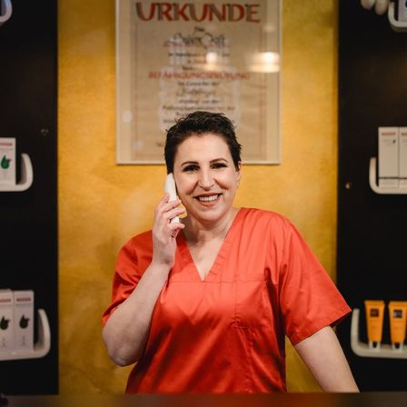 Eine Frau in einem roten Kleid steht an einem Tresen, lächelt und spricht am Telefon in einer Spa-Rezeption. Hinter ihr zeigt eine gelbe Wand ein Zertifikat und Hautpflegeprodukte an.