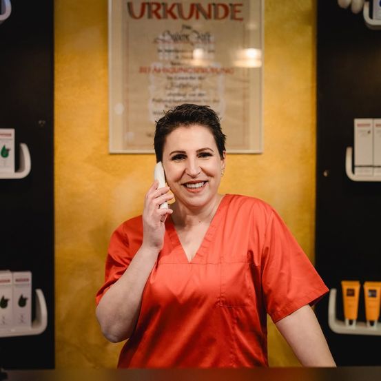 Eine Frau in einem roten Kleid steht an einem Tresen, lächelt und spricht am Telefon in einer Spa-Rezeption. Hinter ihr zeigt eine gelbe Wand ein Zertifikat und Hautpflegeprodukte an.