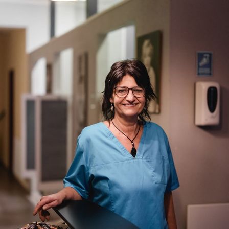 Eine Frau in blauer Arbeitskleidung und mit Brille lächelt in einem Flur mit einem Bild und einem Seifenspender an der Wand.