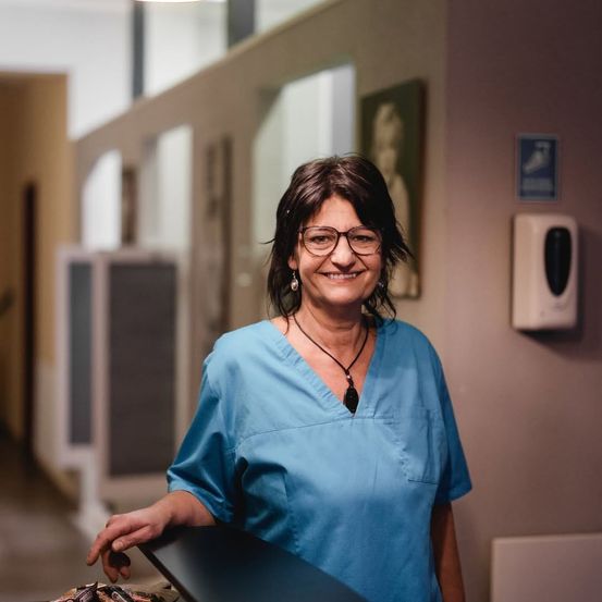 Eine Frau in blauer Arbeitskleidung und mit Brille lächelt in einem Flur mit einem Bild und einem Seifenspender an der Wand.