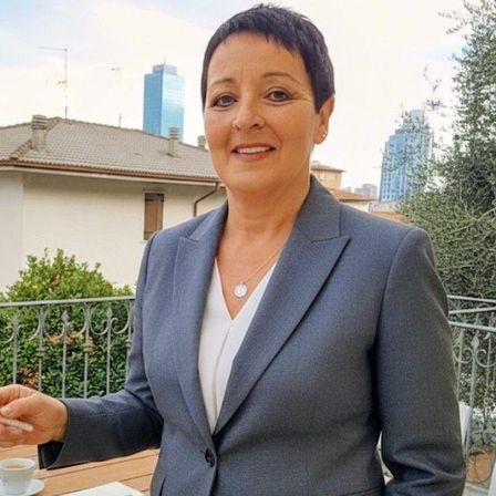 Eine Frau mit kurzen Haaren lächelt für ein Foto, während sie auf einem Balkon steht, eine Zigarette und eine Tasse Kaffee in der Hand hält. Sie trägt einen grauen Blazer und ein weißes Hemd. Hinter ihr sind ein Haus, Pflanzen und entfernte Gebäude zu sehen.
