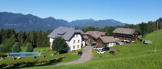 A scenic aerial view of a countryside with a large white house, a swimming pool, and several cars parked in front. The area is surrounded by trees and grass, with mountains in the background.