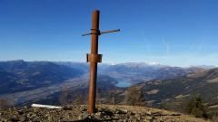Ein Metallkreuz steht auf einem Berggipfel mit einem atemberaubenden Blick auf das Tal und die Berge unter einem klaren blauen Himmel.
