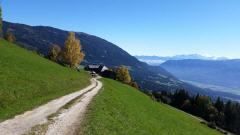 Ein Schotterweg schlängelt sich durch eine saftig grüne Hügellandschaft mit einem entfernten Haus, umgeben von Bergen und einem klaren blauen Himmel.