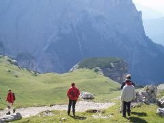 Fünf Personen wandern auf einem grasbewachsenen Bergpfad mit einem felsigen Felsvorsprung und einer großen Bergkette im Hintergrund.
