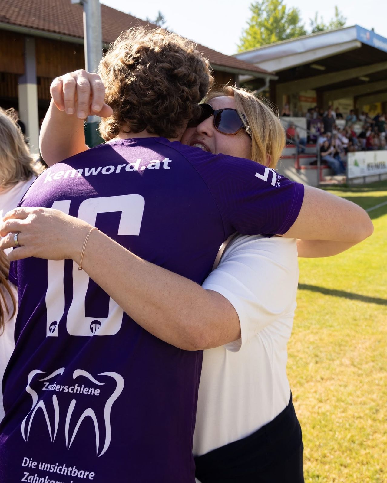 A man wearing a purple shirt hugs a woman wearing a white shirt. They are standing on a field in front of a building.