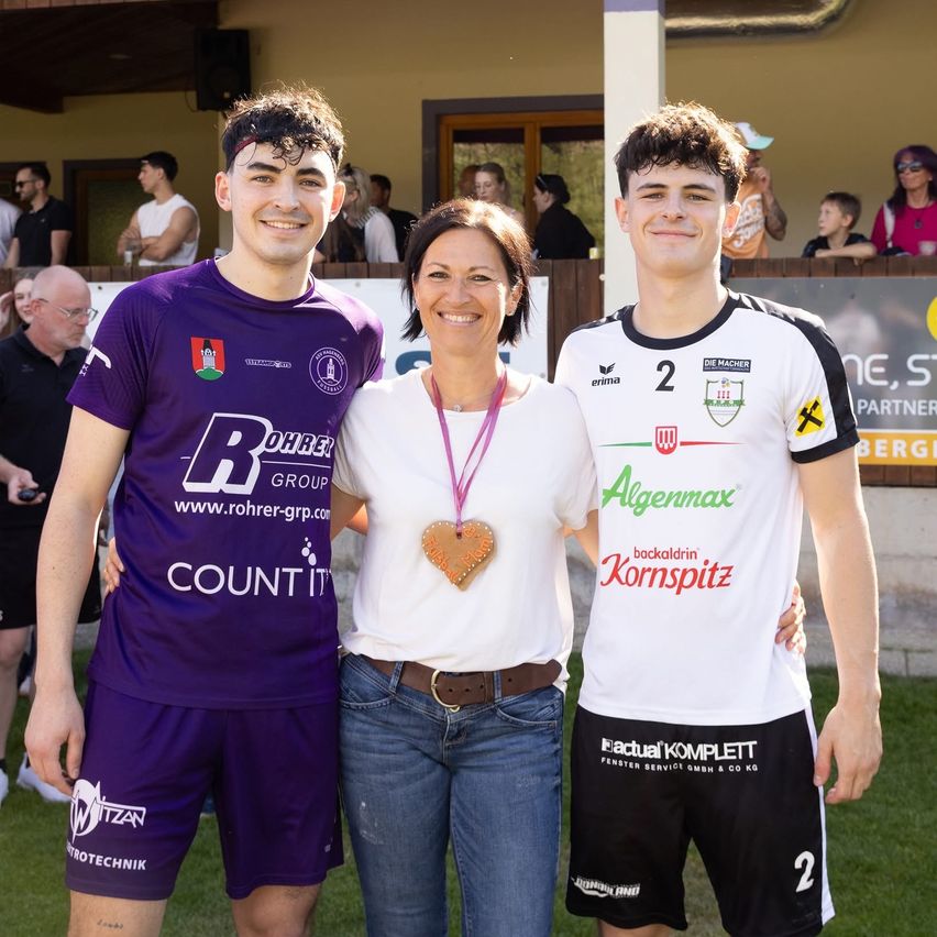Three individuals pose for a photograph in an outdoor setting, with two young men wearing sports jerseys and a woman wearing a white shirt. Behind them, there are people standing and watching in the background.