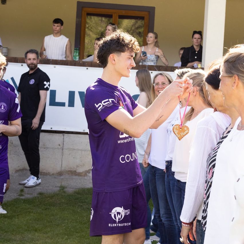 A soccer player in a purple jersey hands a medal to a woman while others watch from behind a barrier with the word 'Liv'.