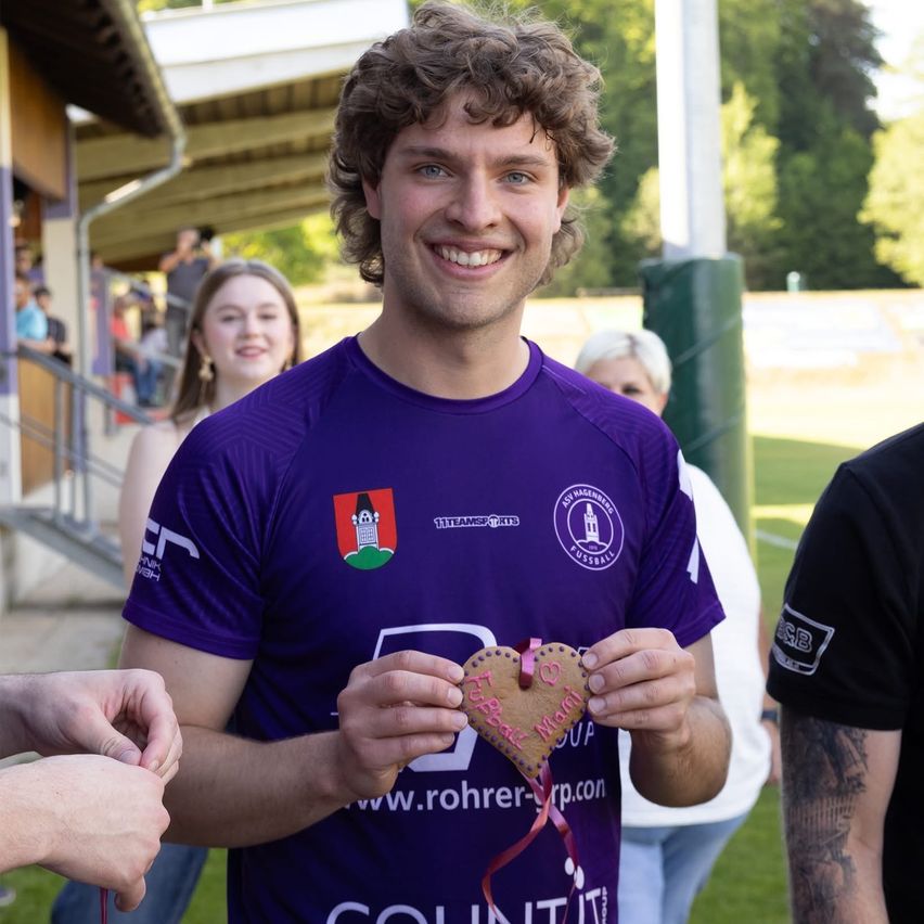 A man in a purple shirt holds a heart-shaped cookie, smiling in an outdoor setting with others in the background.