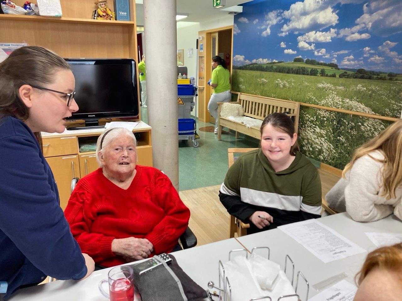 Eine Frau in einem roten Pullover sitzt an einem Tisch mit einem jungen Mädchen in einer grün-schwarzen Kapuzenjacke. Sie lächeln beide, eine Frau in einem grünen Shirt putzt in der Nähe.