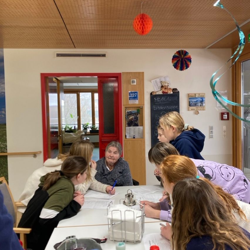 Eine Gruppe von Mädchen sitzt mit einer älteren Frau um einen Tisch herum, sie scheinen etwas zu besprechen. Der Raum hat eine weiße Wand, eine Tafel und Dekorationen. Der Raum ist gut beleuchtet, mit Sonnenlicht, das durch die Fenster scheint.