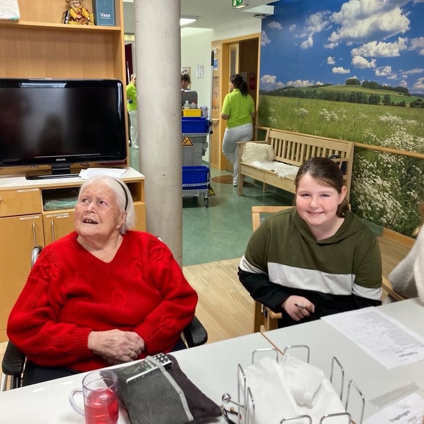 Eine ältere Frau in einem roten Pullover sitzt in einem Rollstuhl mit einer Fernbedienung neben ihr. Ein junges Mädchen in einer grünen und weißen Kapuzenjacke sitzt neben ihr. Hinter ihnen steht ein Schreibtisch mit Papieren, einem Glas Saft und einer Fernbedienungstasche.