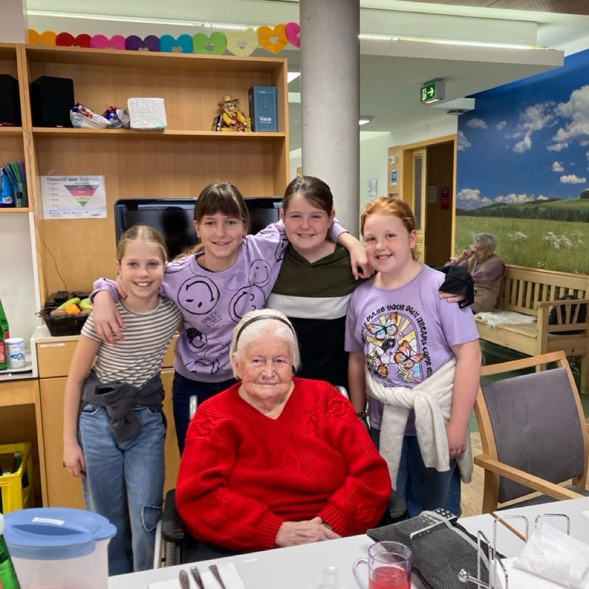 Vier Mädchen in passenden lila Shirts stehen hinter einer älteren Frau im Rollstuhl und posieren für ein Foto in einem Raum mit einem Wandbild und einer Bank.