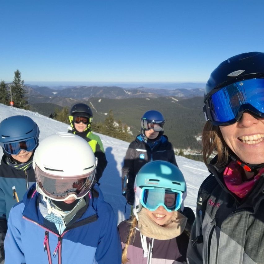 Fünf Personen stehen auf einem verschneiten Berg. Sie tragen Skiausrüstung, Helme und Schutzbrillen. Dahinter sind ein Baum und Berge. Der Himmel ist blau.