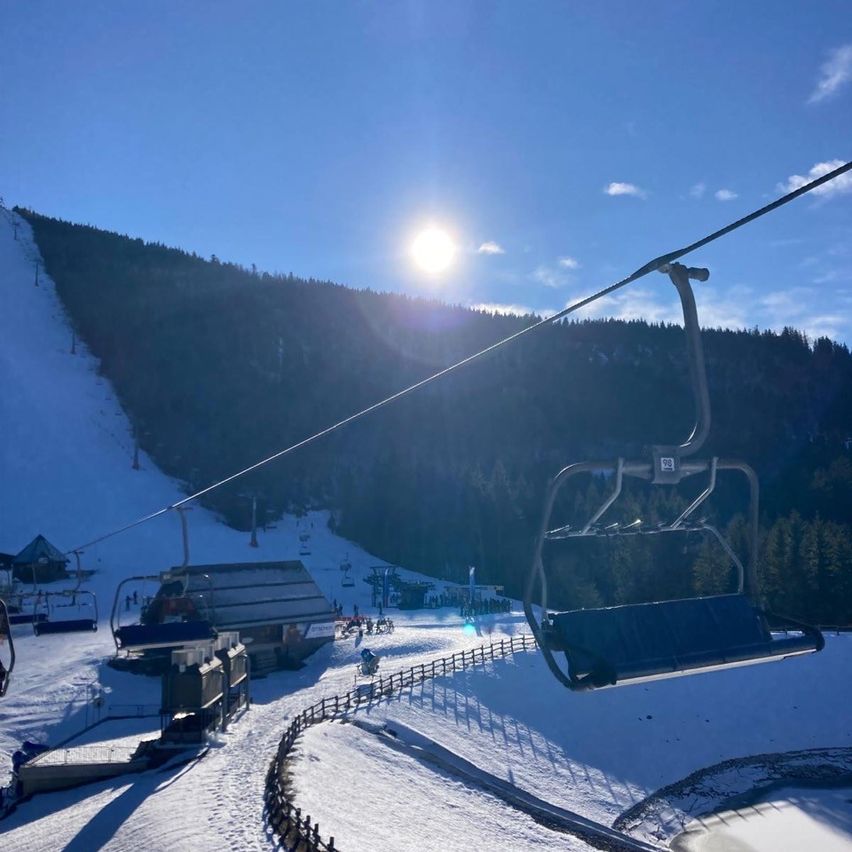 Eine Skilift in einem verschneiten Gebirgsgebiet unter einem strahlenden Sonnenschein, mit einer Piste und mehreren Personen im Hintergrund.