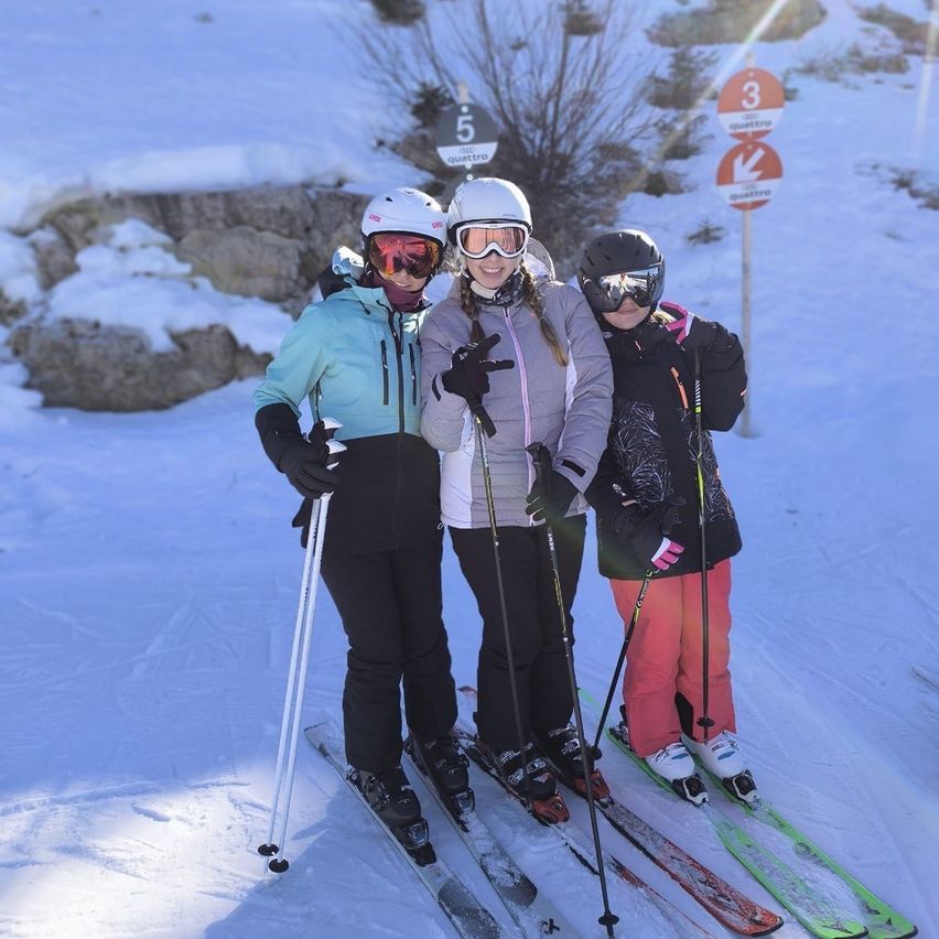 Drei Personen posieren für ein Foto auf einem verschneiten Berg. Sie tragen Winterkleidung und Skiausrüstung. Hinter ihnen befindet sich ein Schild mit Zahlen und Pfeilen.