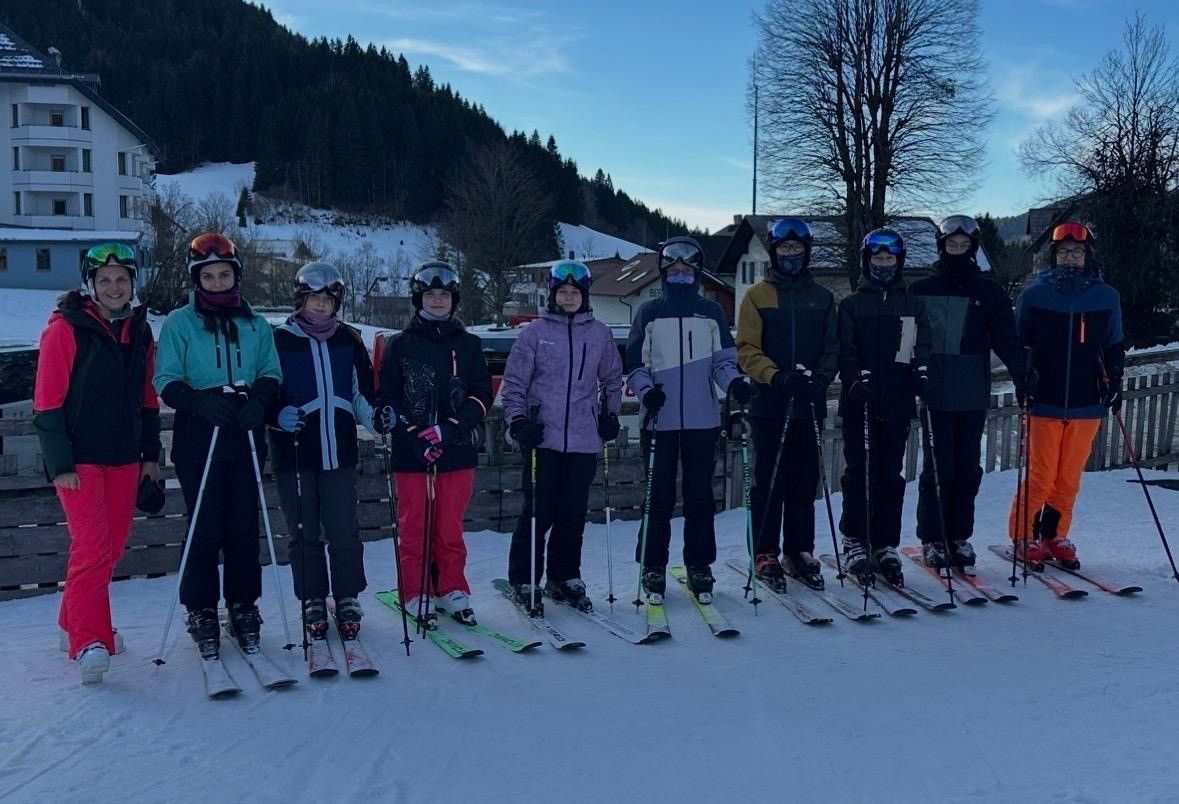 Eine Gruppe von Skifahrern posiert für ein Foto auf einem verschneiten Berg und trägt Winterkleidung. Der Hintergrund zeigt Bäume, ein Gebäude und einen klaren blauen Himmel.