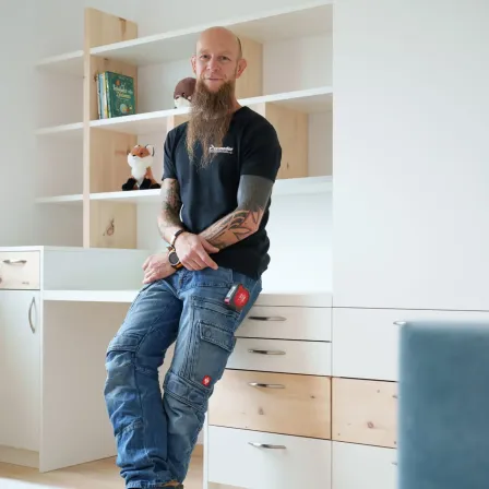 Ein Mann mit Bart und Tattoos sitzt auf einem Schreibtisch in einem Raum mit weißen Wänden und Holzregalen.