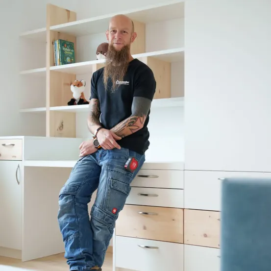 Ein Mann mit Bart und Tattoos sitzt auf einem Schreibtisch in einem Raum mit weißen Wänden und Holzregalen.