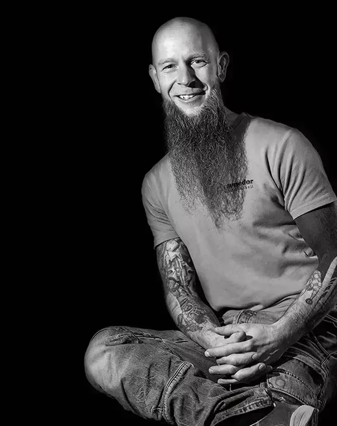 Ein Schwarz-Weiß-Porträt eines kahlköpfigen Mannes mit langem Bart und Tattoos, der mit einem Lächeln auf dem Gesicht sitzt.