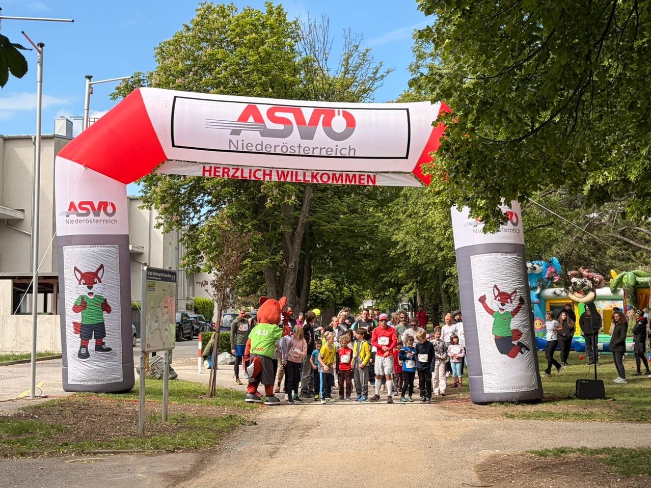 Ein roter Fuchsmascot heißt eine Gruppe von Kindern in grünen und roten Shirts unter einem ASVÖ Niederösterreich-Bogen willkommen, mit Bäumen und einem Gebäude im Hintergrund.